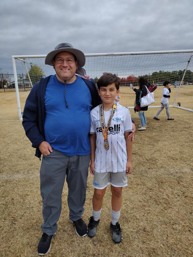 Chris Platania, Team Member of the Year, stands with son in front of soccer goal.

