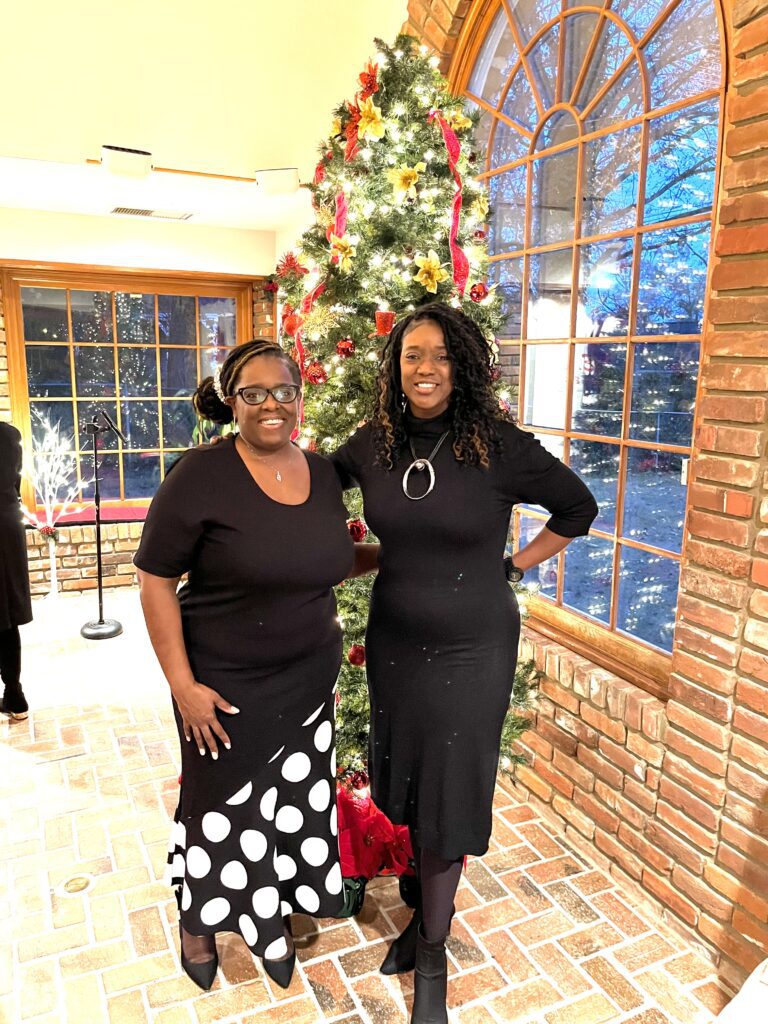 Two women in black dresses stand in front of a Christmas Tree
